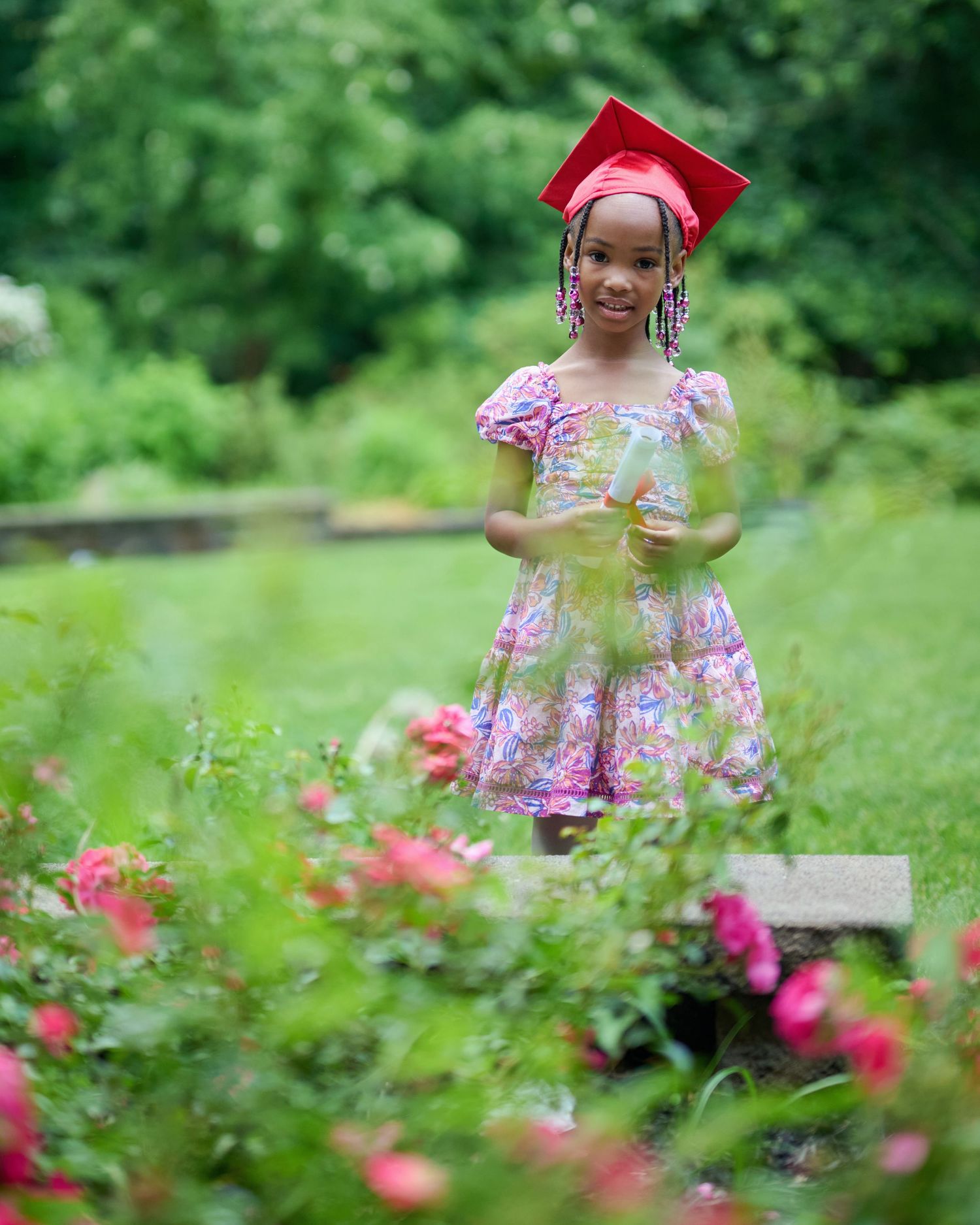 Garden graduation photos with pink floral dress and red cap among blooming flowers.