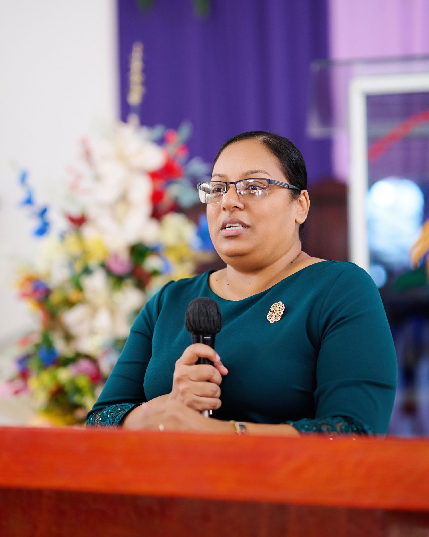 Speaker in dark attire presenting at wooden podium with floral arrangements.