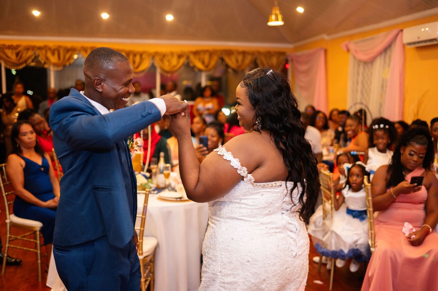 Groom in blue suit kisses bride's hand at wedding reception while guests watch in decorated venue.