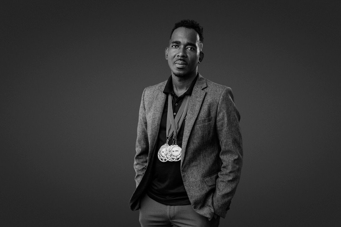 Black and white portrait of a young man in a blazer wearing a decorative medallion necklace, posed confidently.