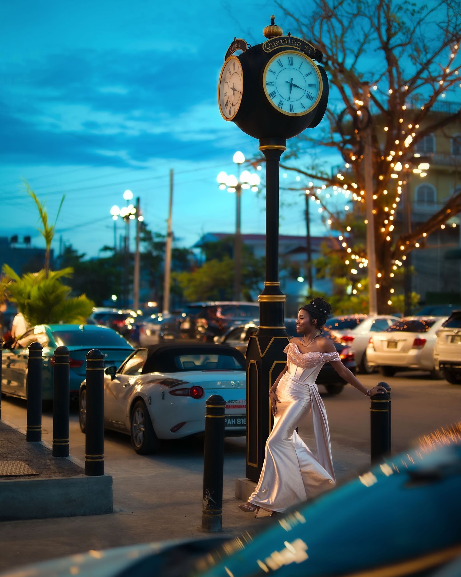 Woman in white gown poses by ornate street clock at twilight, surrounded by cars and twinkling lights.