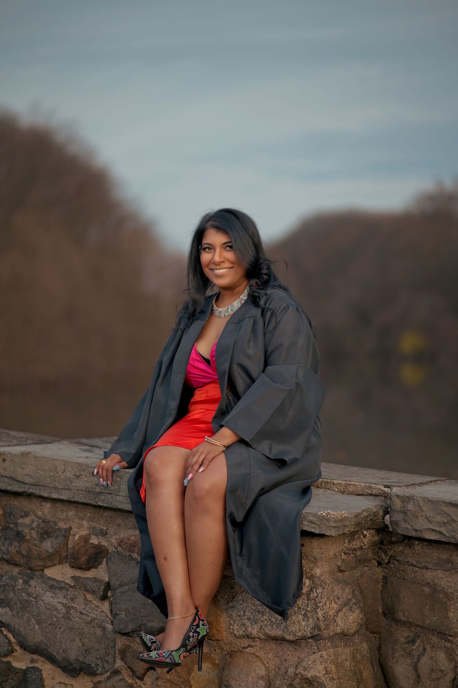A graduate in black regalia poses on a stone wall during a sunset photoshoot.