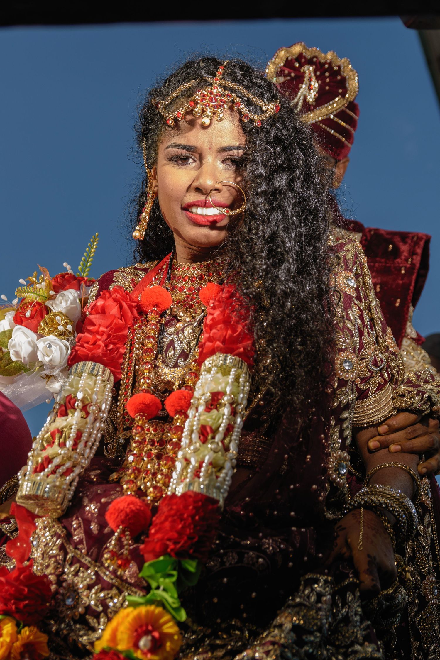 A bride in an ornate red and gold traditional Indian wedding dress holds a colorful bouquet.