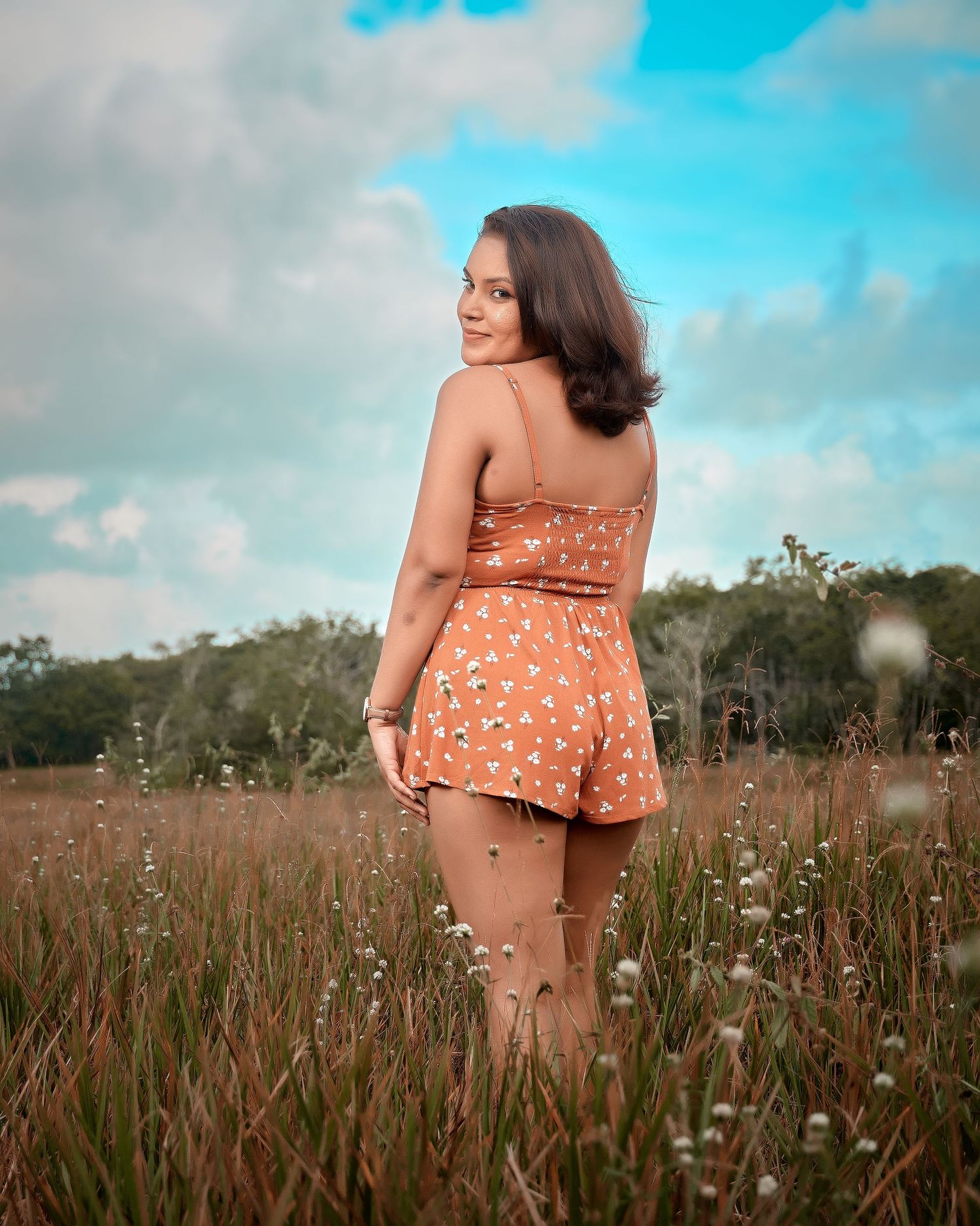 Person wearing orange floral dress stands among tall grass in a natural outdoor setting with blue sky.