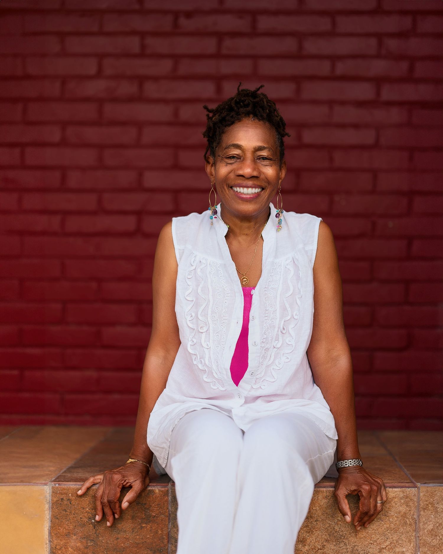 A joyful portrait against a red brick wall wearing a white sleeveless dress with pink accent.