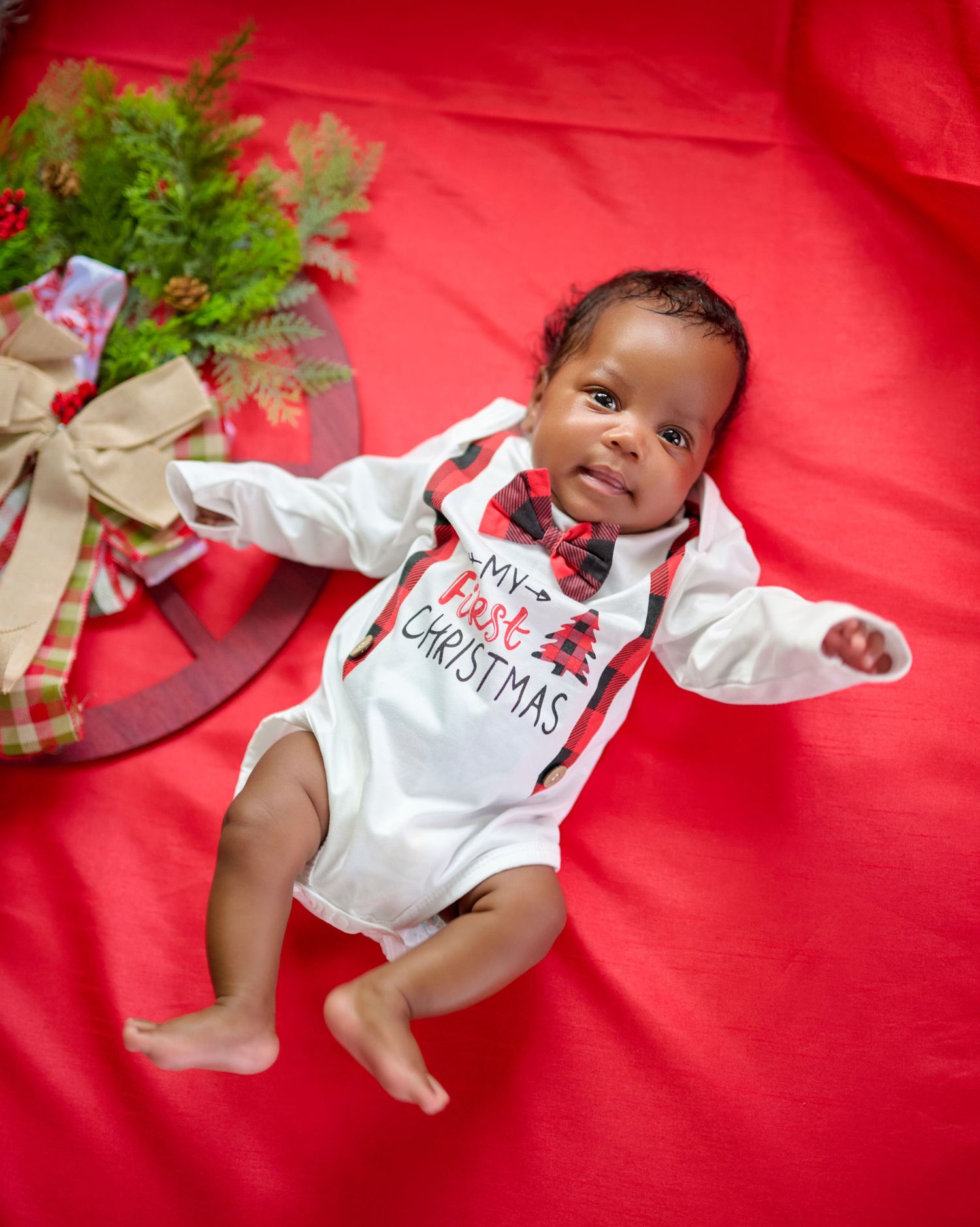 Holiday themed photo of baby wearing white onesie on red backdrop with Christmas decorations.