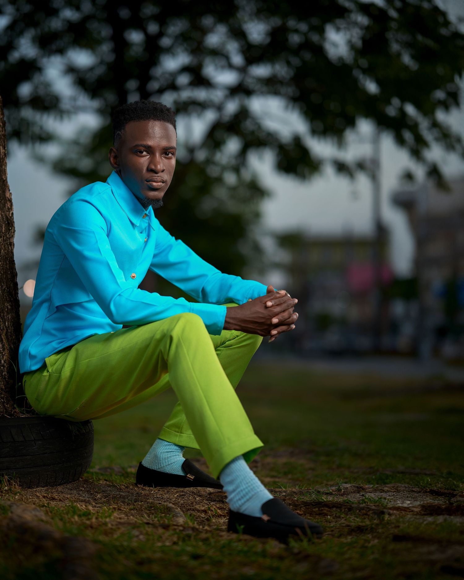 Casual outdoor pose in vibrant blue shirt and neon yellow pants on grassy ground.