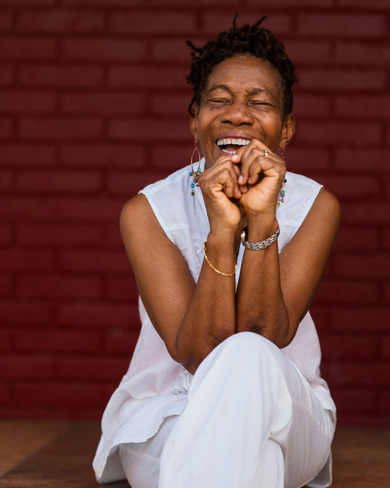 A person in white clothing laughs joyfully against a red brick wall background.