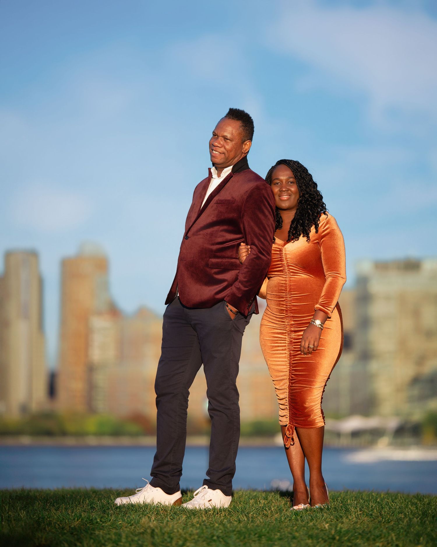 A couple in formal attire poses together on a grassy hill with city buildings in the background.