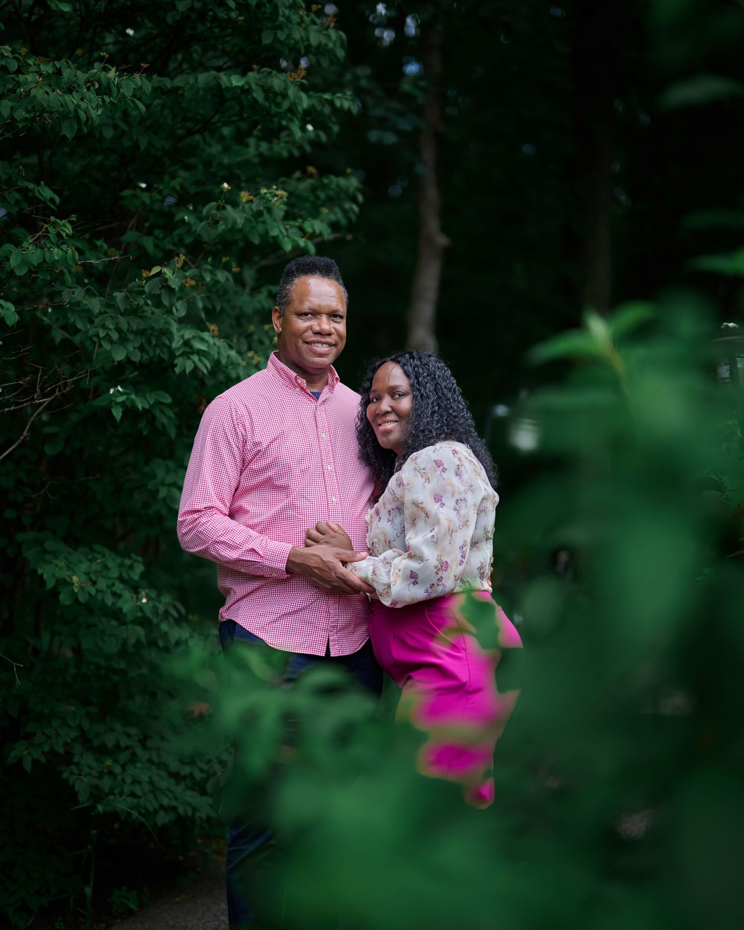 Couple in pink attire photographed through foliage in a forest setting.