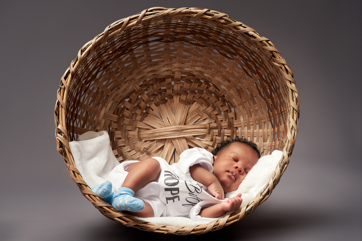 Overhead view of sleeping newborn in round wicker basket against gray background.