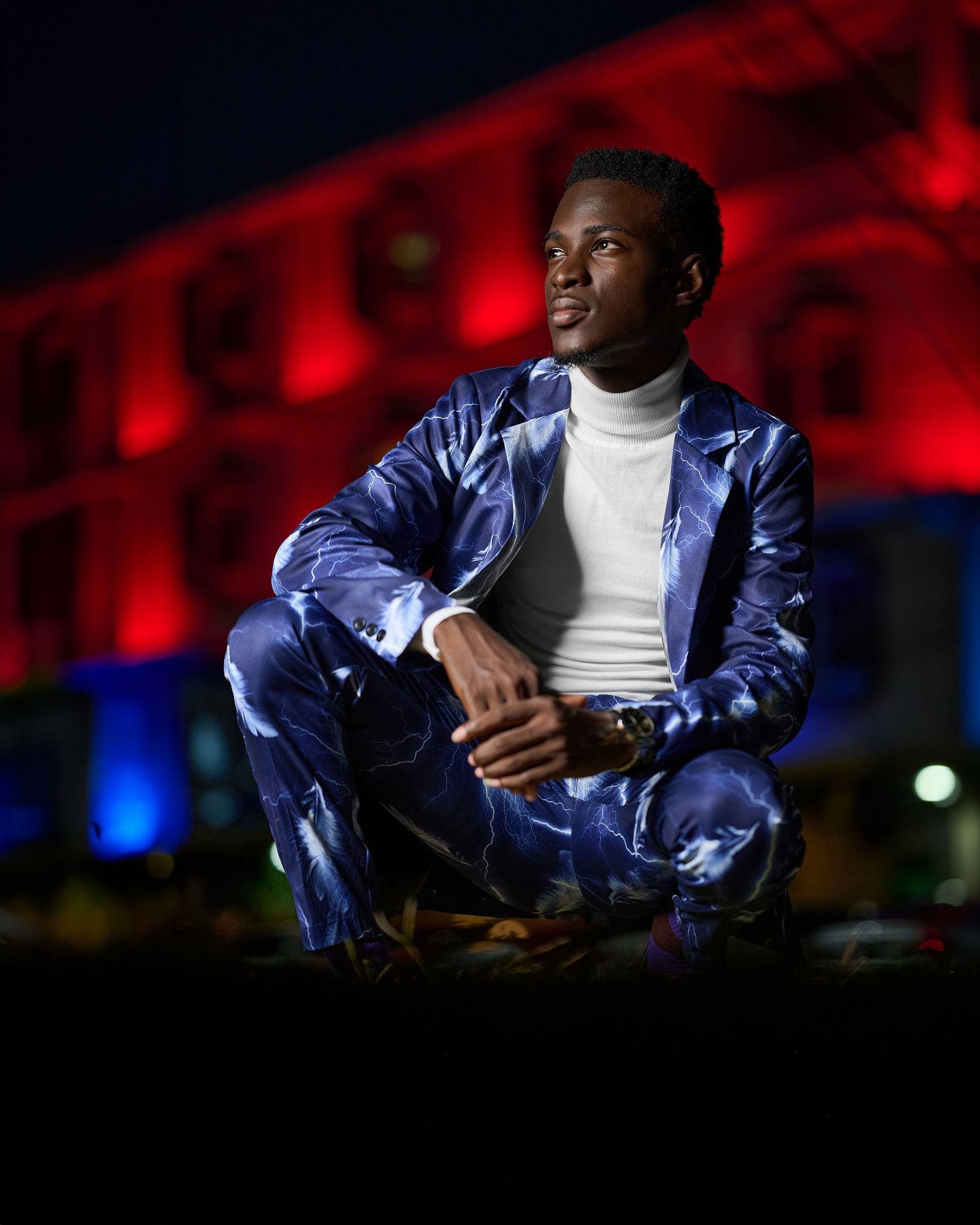 Young man in blue satin suit and white turtleneck sits confidently outdoors at night with red-lit building backdrop.