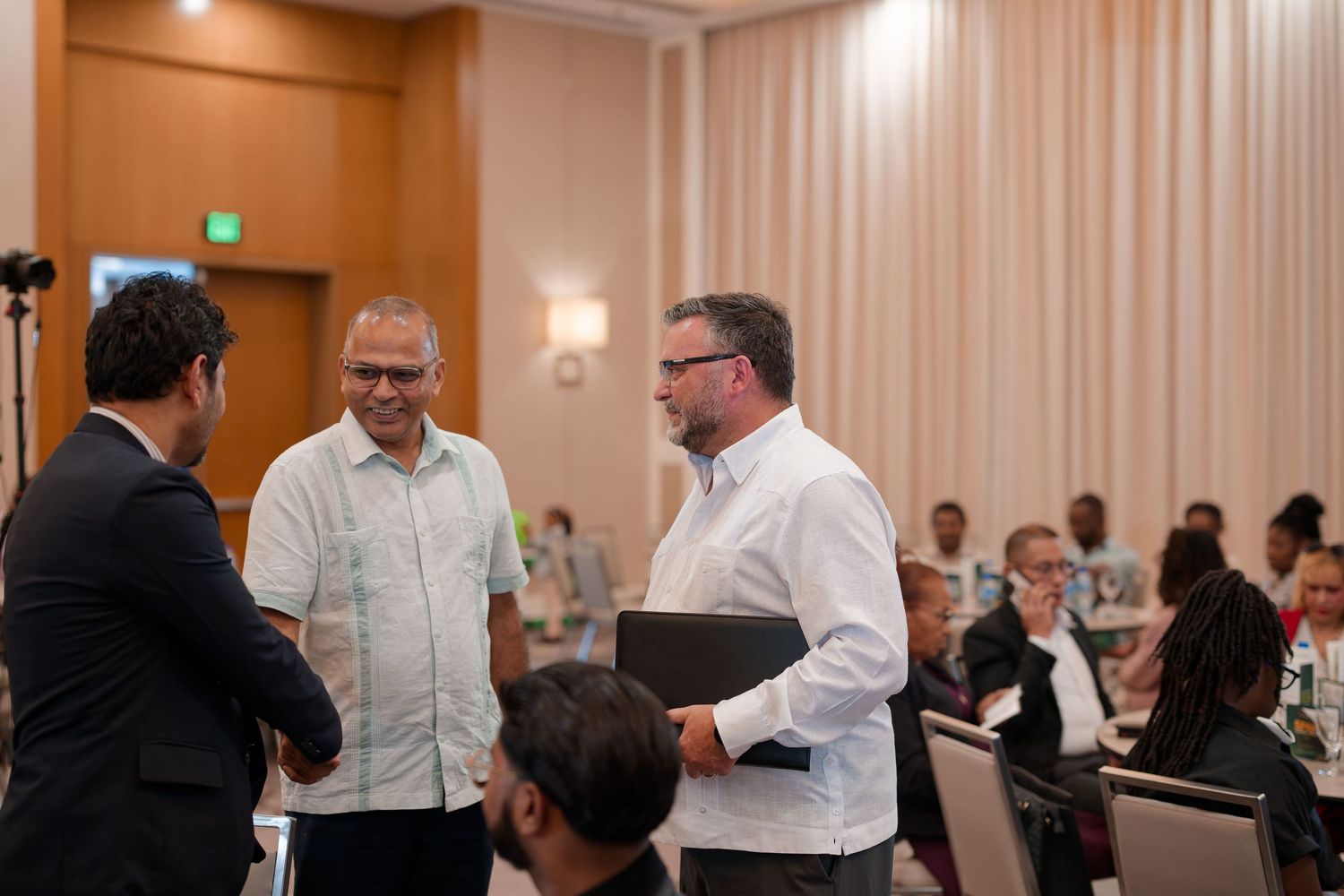 Three professionals networking and shaking hands at a formal conference event in a hotel ballroom.