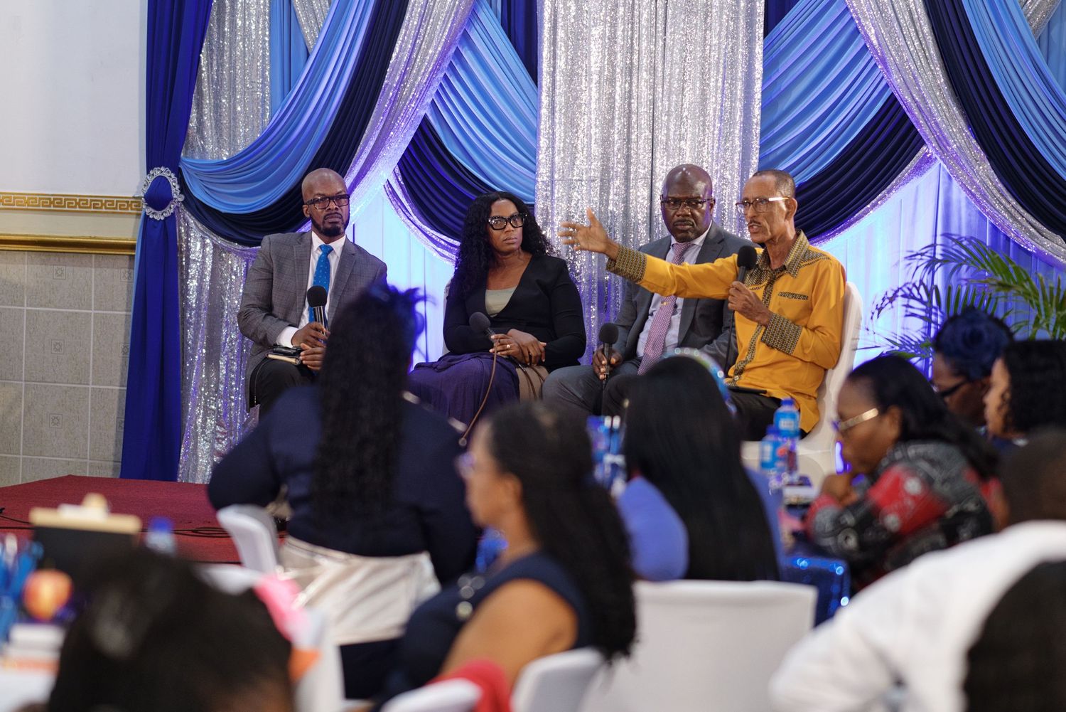 Panel discussion with four speakers seated on stage before an audience at a formal event with blue drapes.