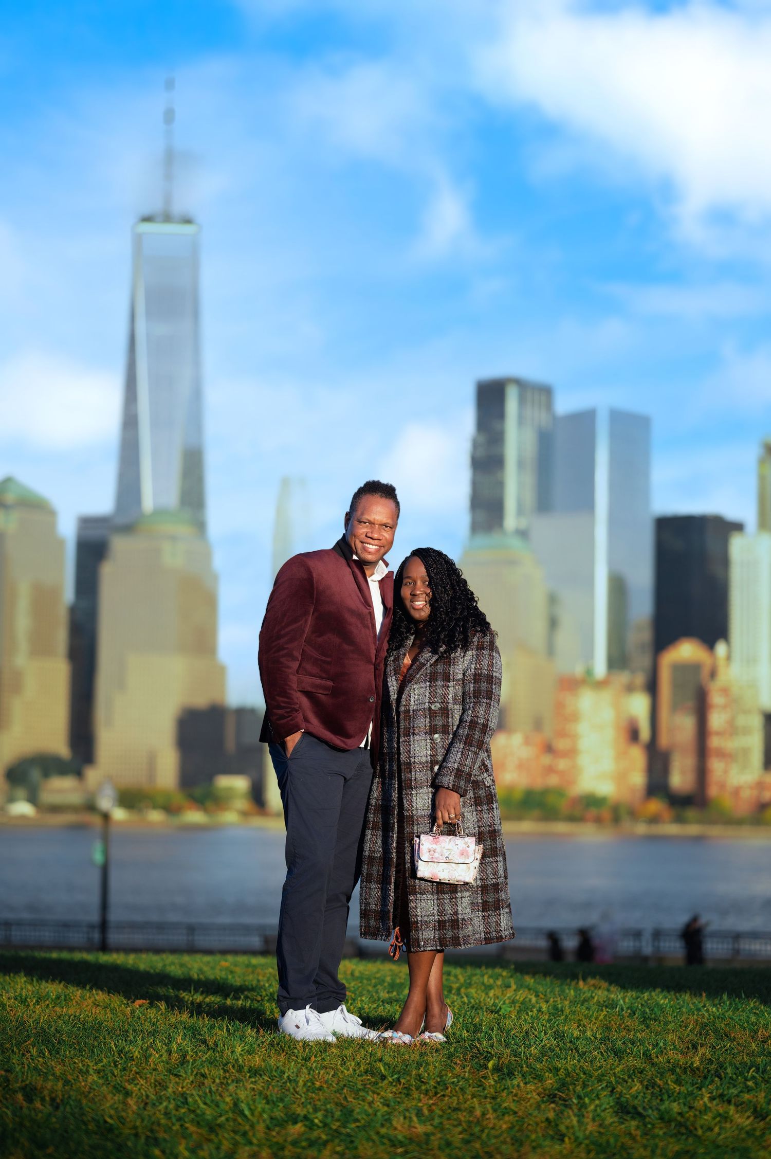 A couple stands on grass with the New York City skyline visible in the background.