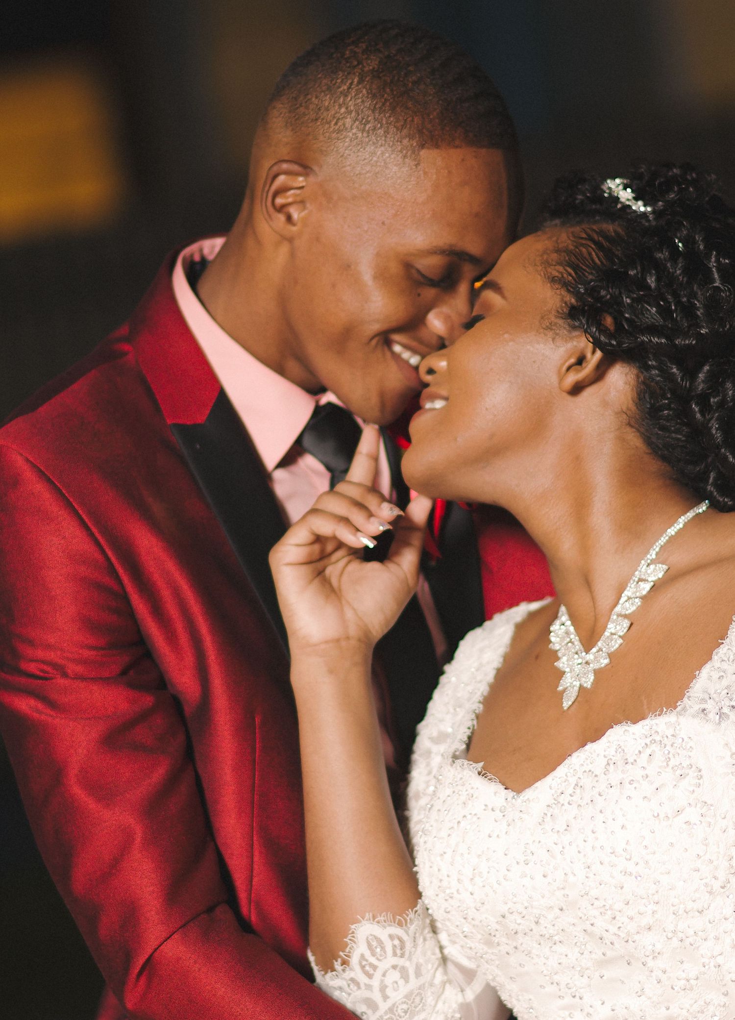 A bride and groom share a romantic moment, the groom in a red blazer and bride in white lace dress with crystal necklace.