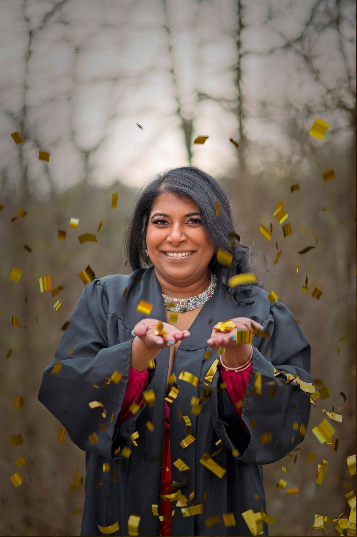 Graduate celebrating with gold confetti toss against winter trees background.