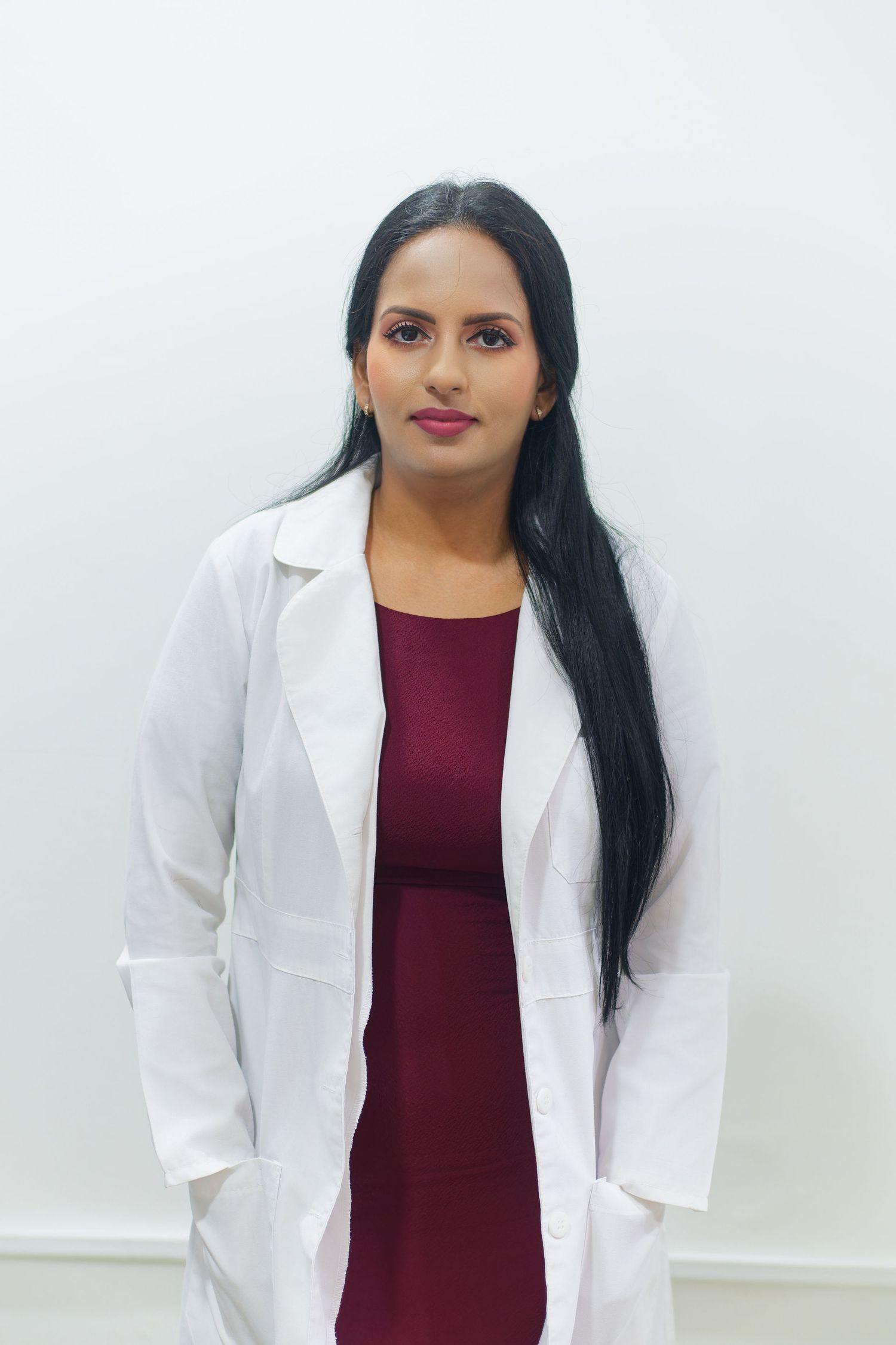 Healthcare professional in white coat and burgundy dress standing against white background.