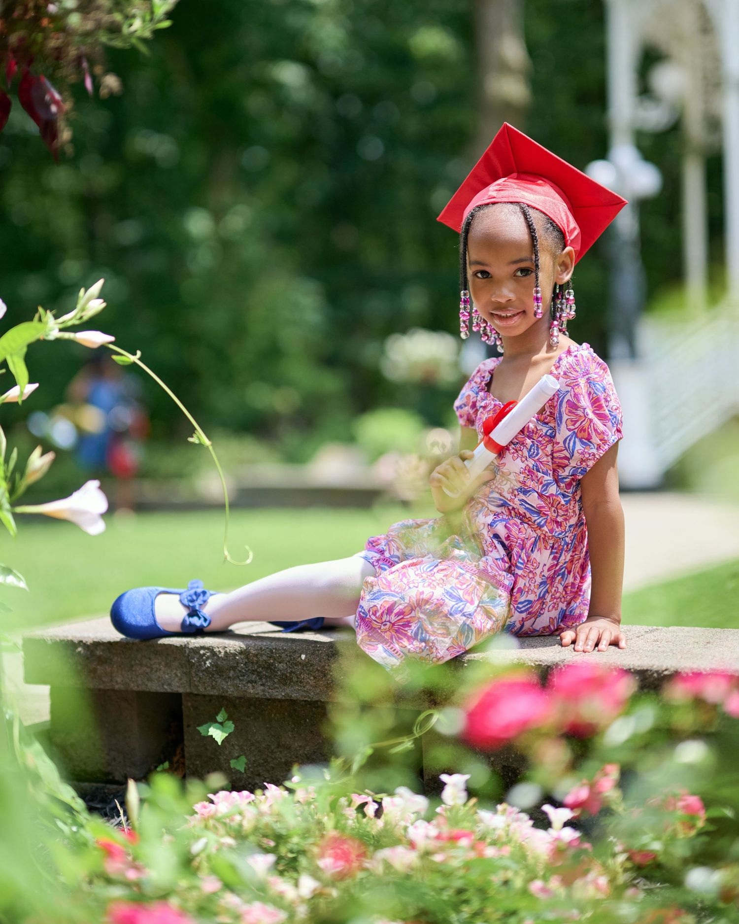 Graduate in floral dress sits on stone wall surrounded by pink roses in garden setting.