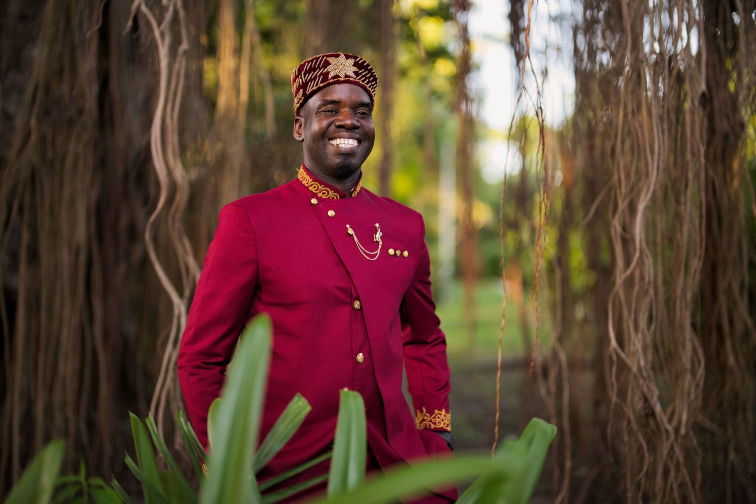 Smiling man in red African traditional attire and kufi hat standing among trees in a lush green setting.