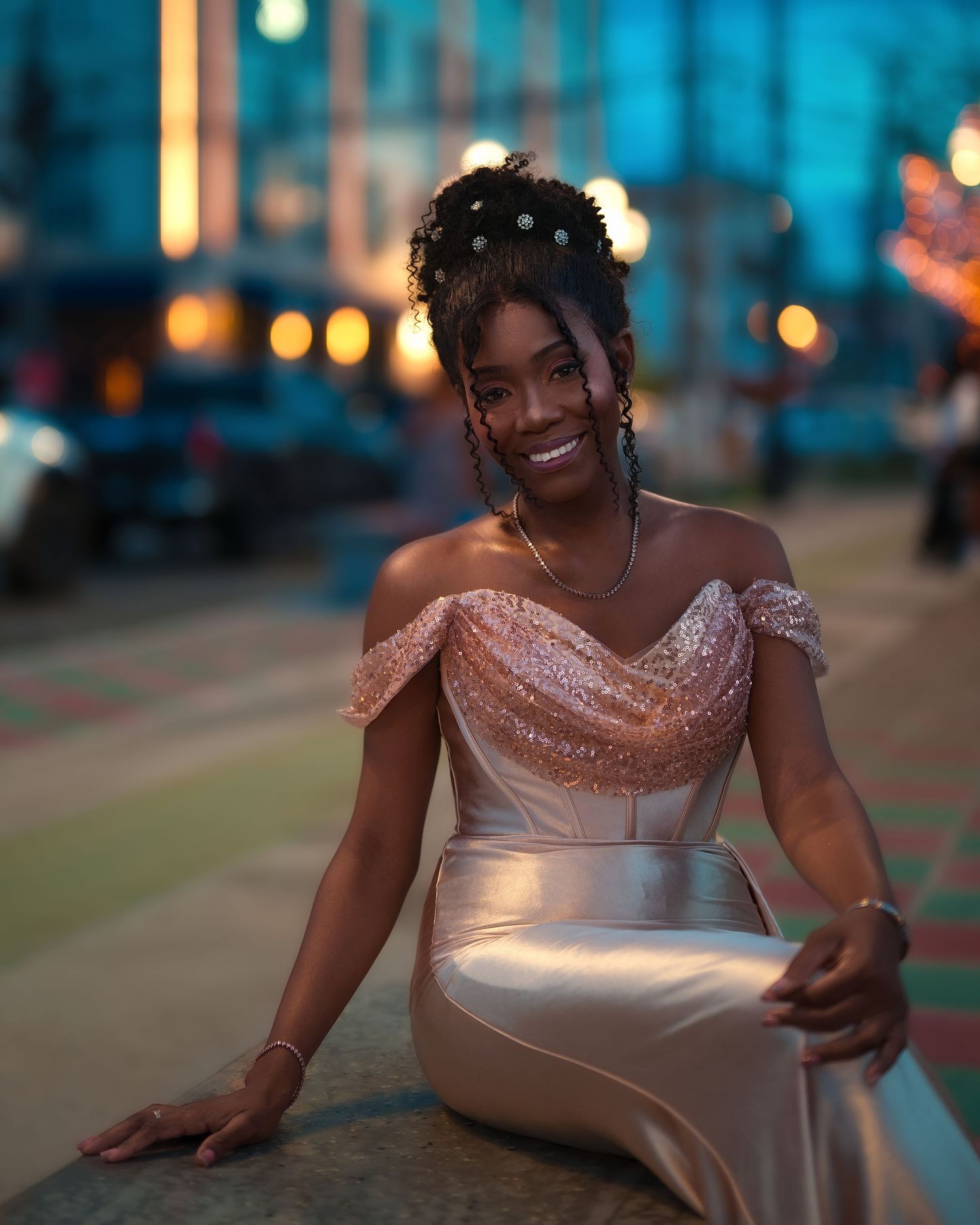 A smiling woman in a elegant off-shoulder sequin gown sits outdoors at dusk with bokeh city lights behind her.