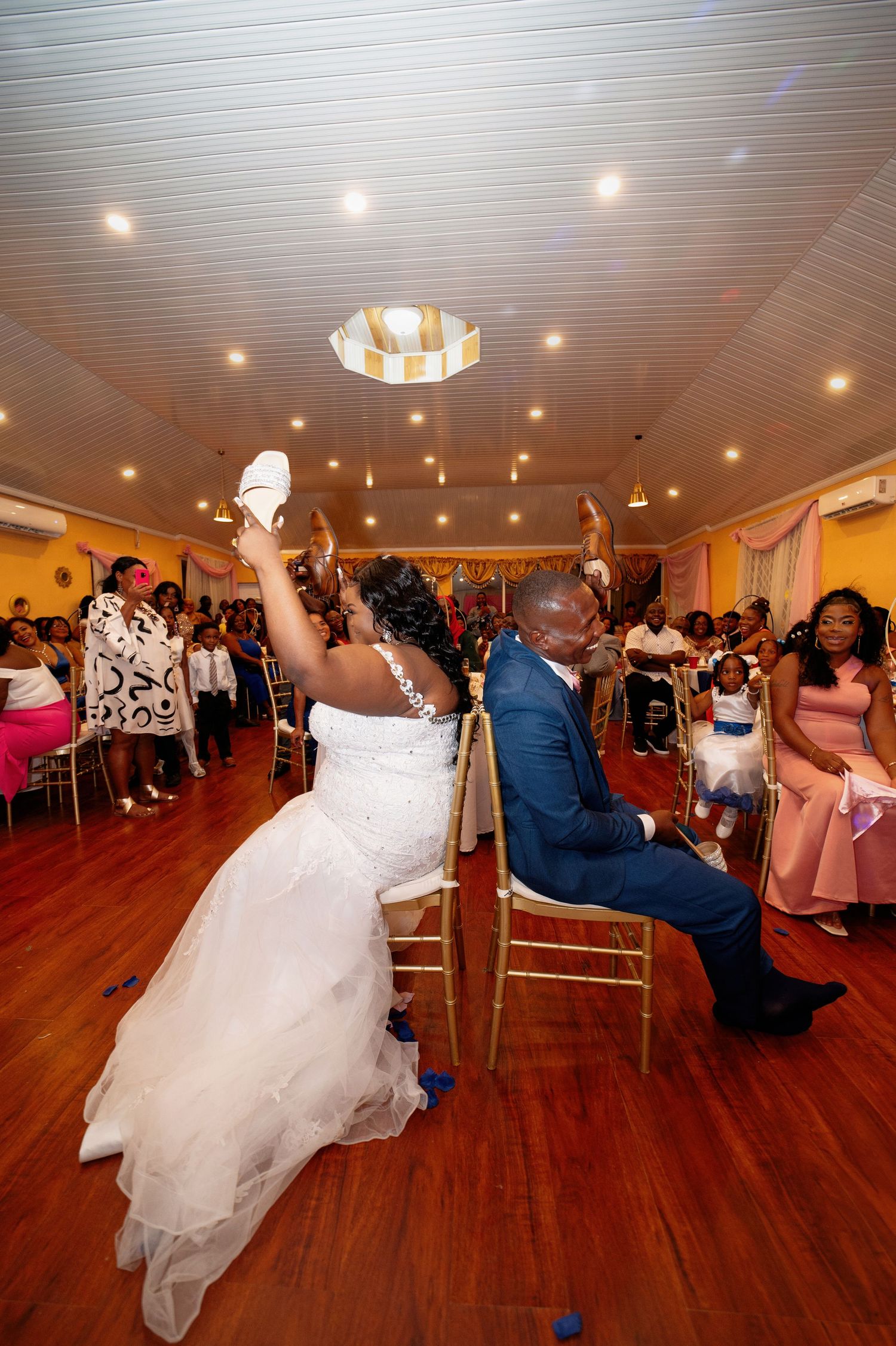 Bride and groom celebrate at wedding reception, bride tossing shoe in air while guests cheer in decorated banquet hall.