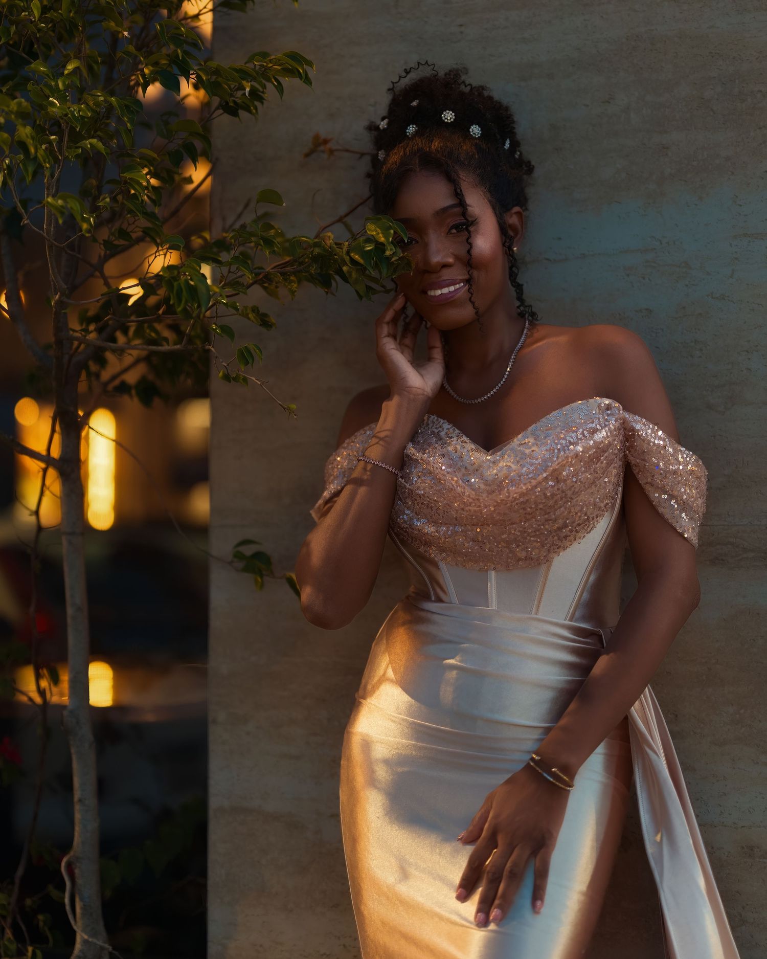 A smiling woman in an elegant off-shoulder sequined wedding gown with pearl hair accessories poses outdoors at dusk.