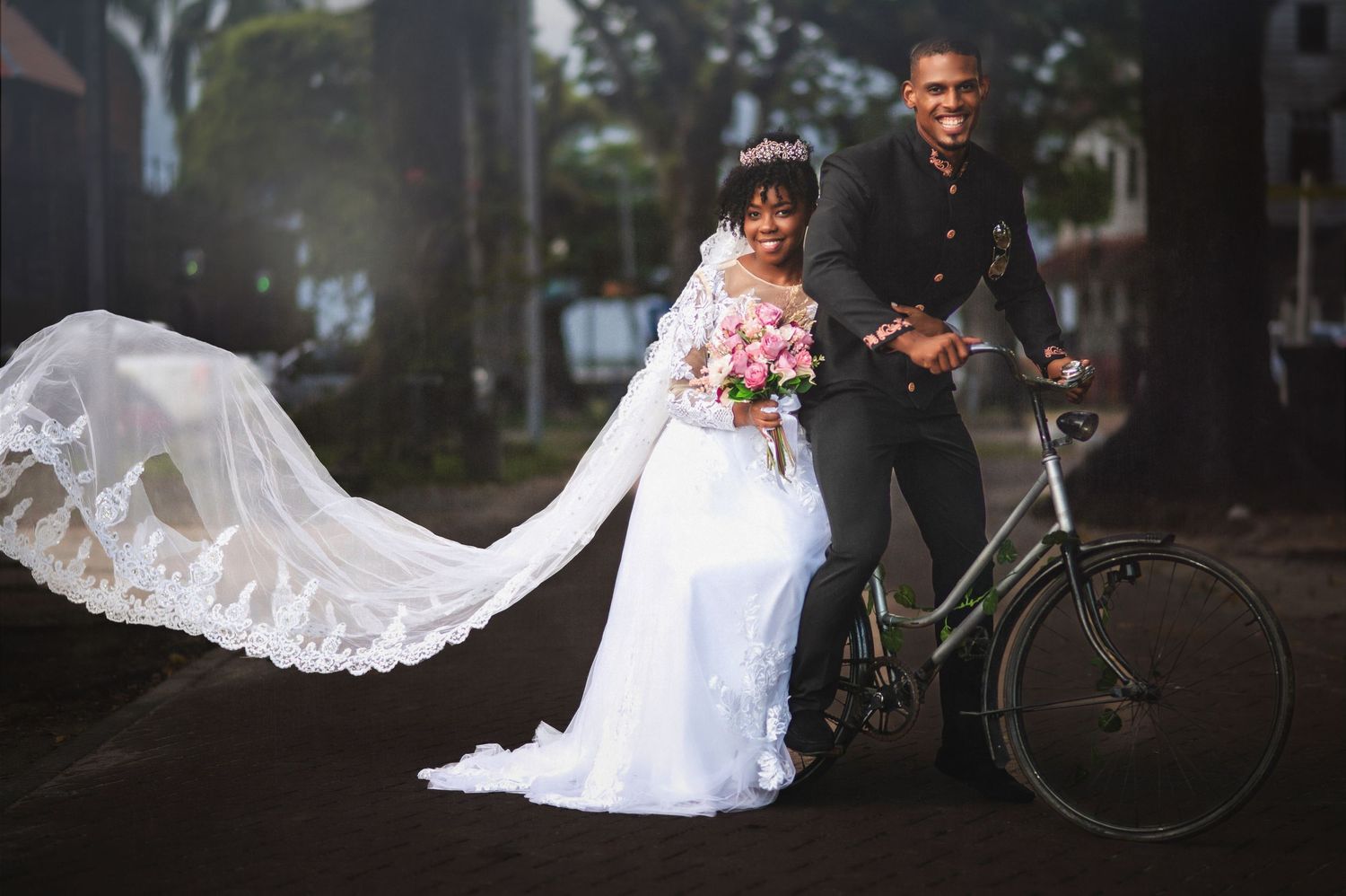A bride in a white wedding dress poses with a vintage bicycle in an outdoor garden setting.