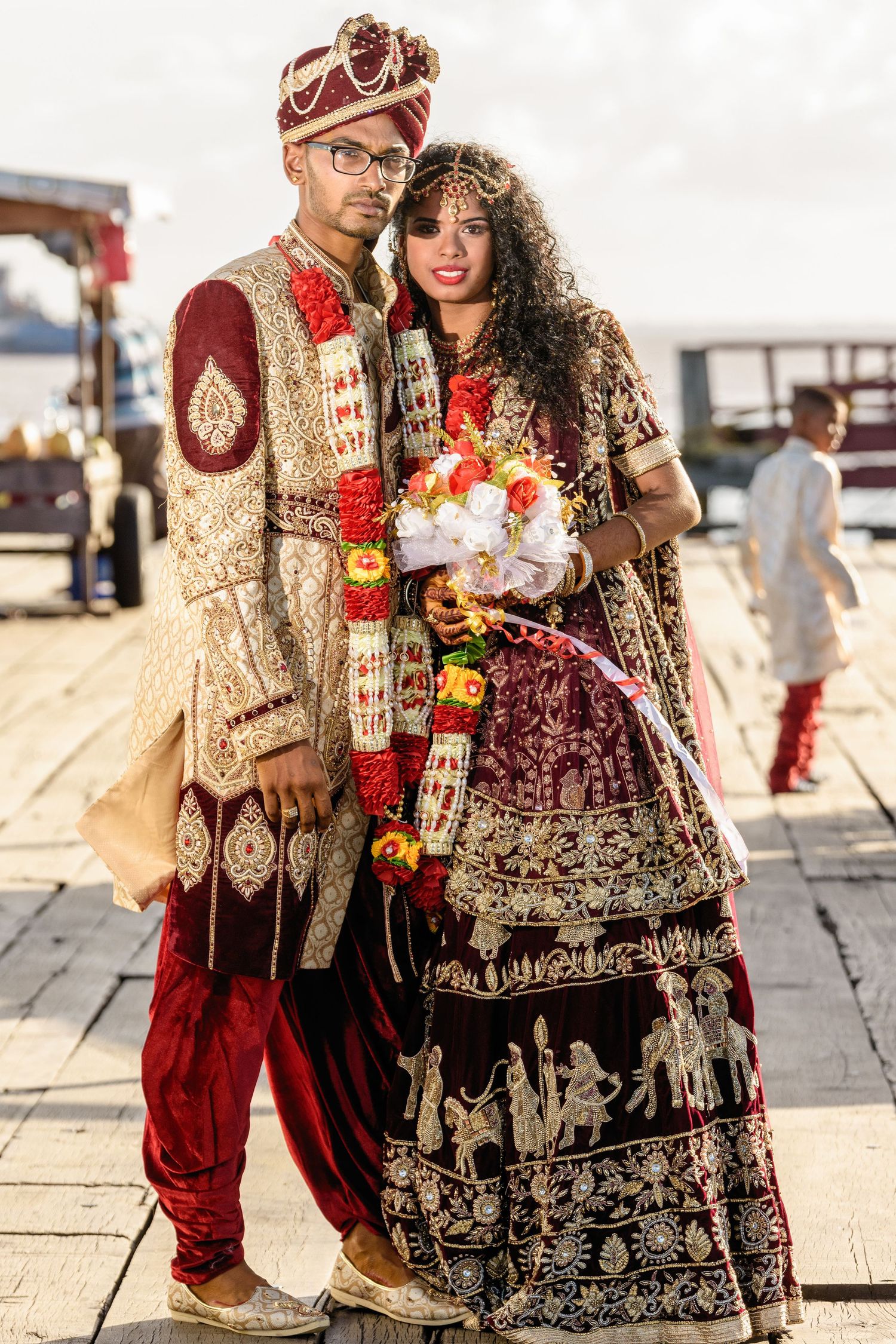 A couple in traditional Indian wedding attire poses together outdoors, wearing ornate red and gold garments.