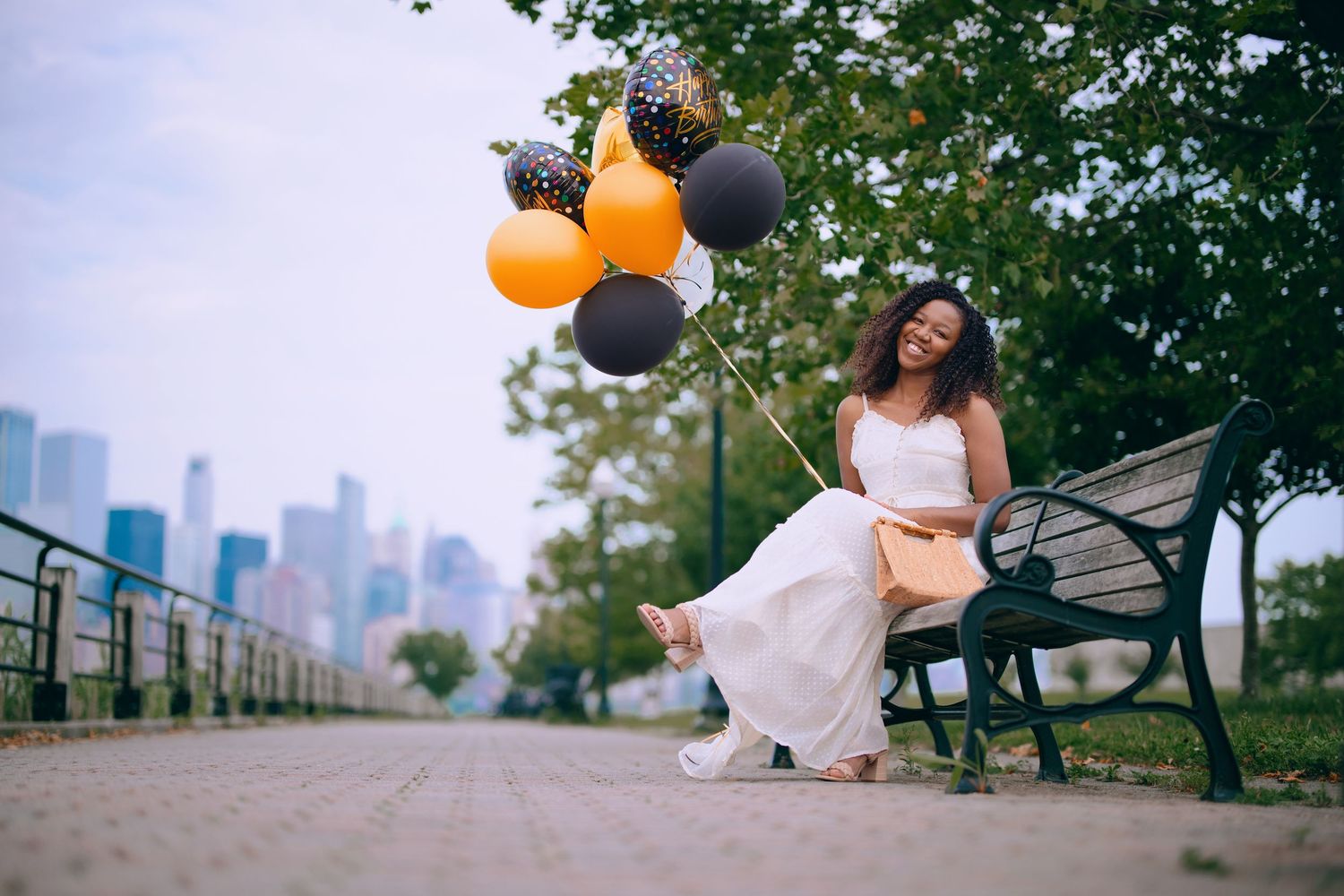 Person in white dress poses with balloons on grassy hill overlooking urban skyline at sunset.