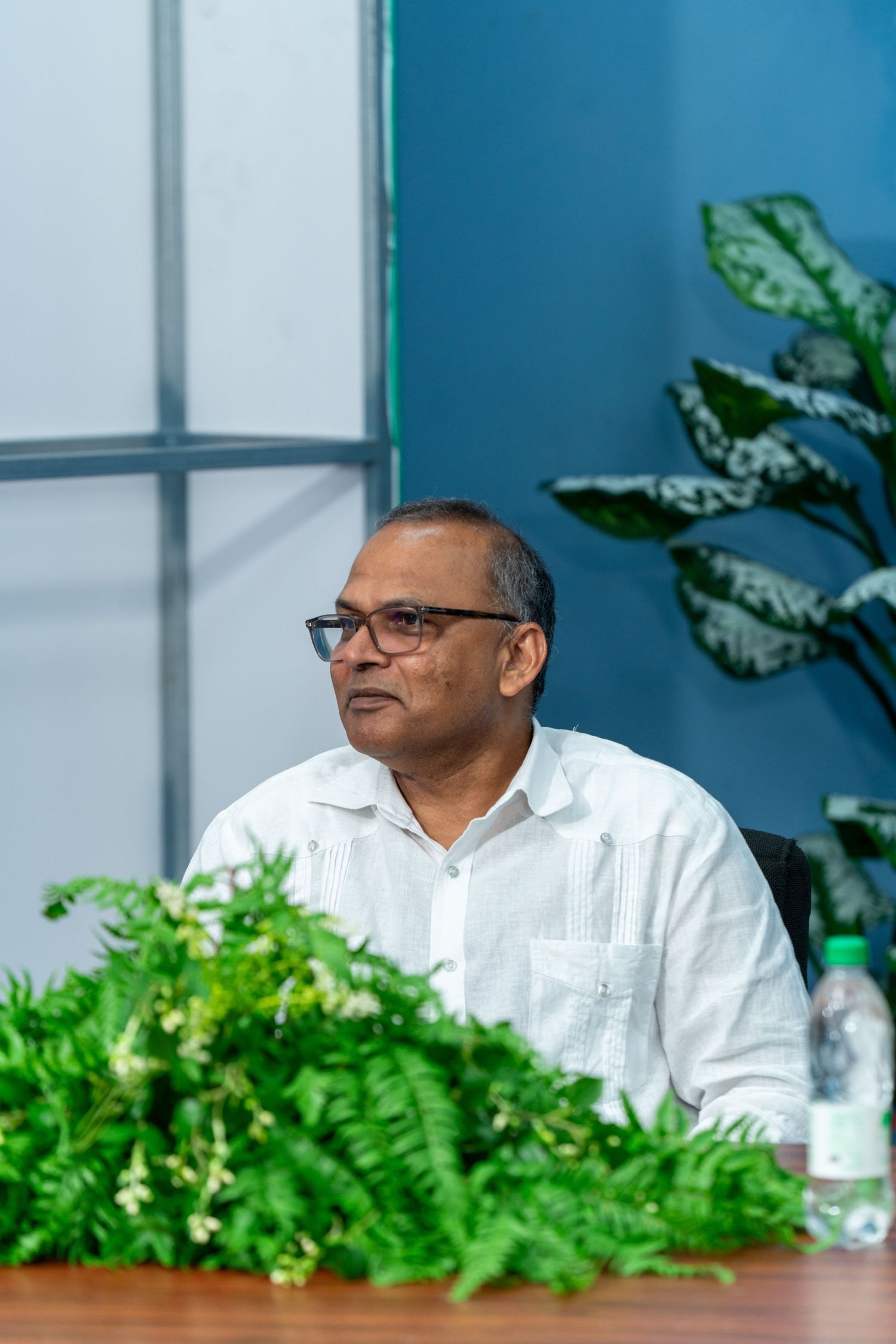 Speaker in white dress shirt seated at conference table with green plants in background.