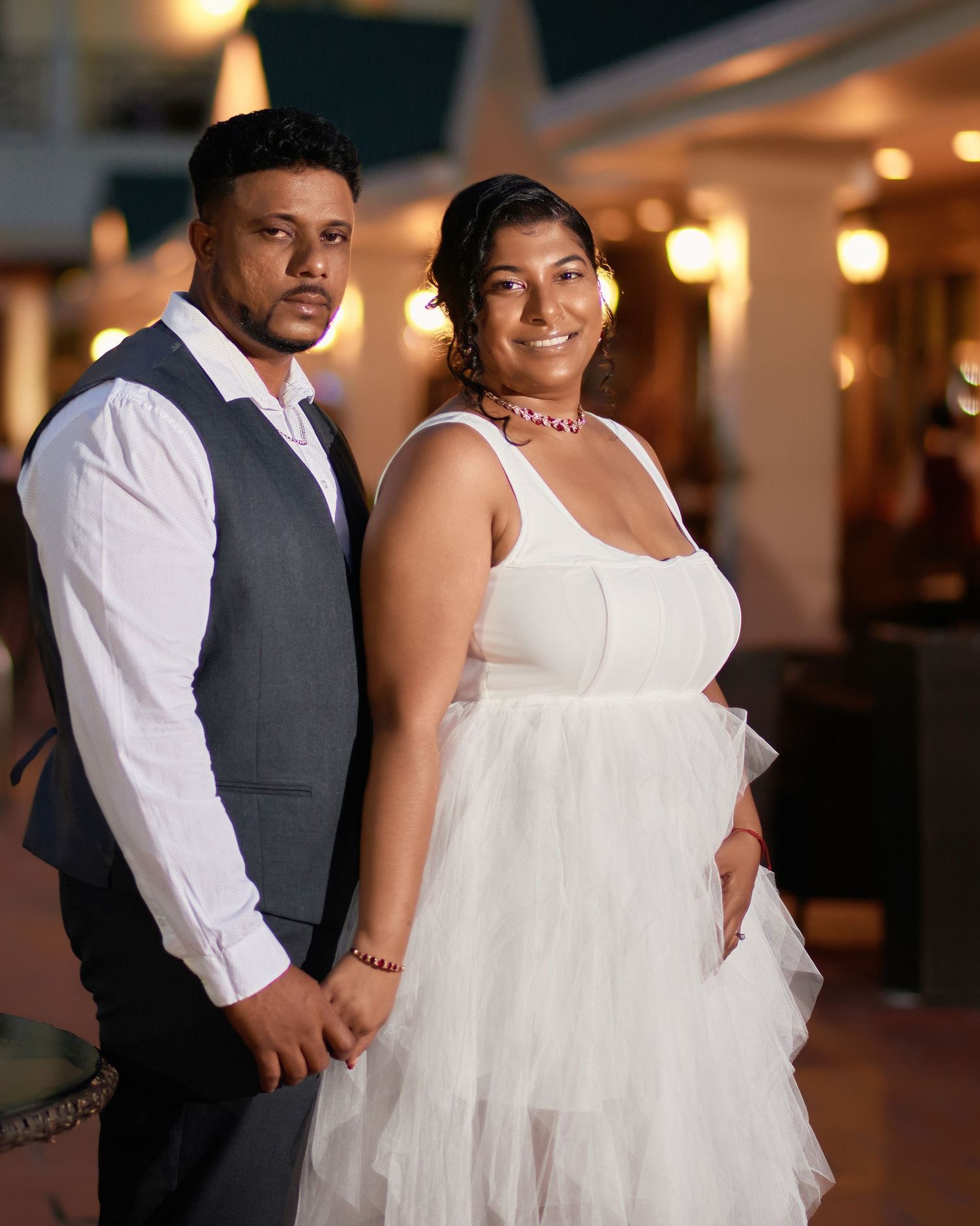 A couple in formal white attire poses together at an indoor celebration venue.