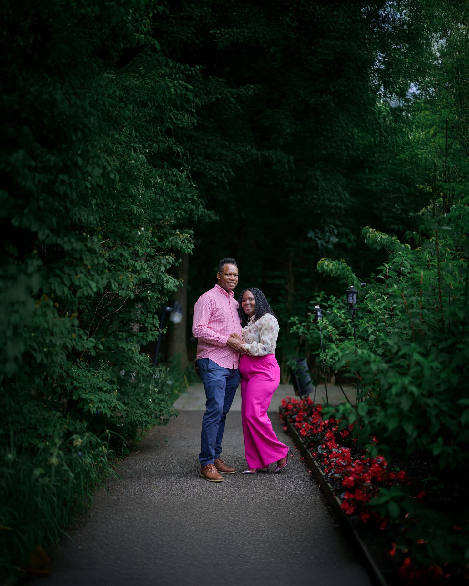 A couple poses together in a garden path surrounded by lush greenery and red flowers.