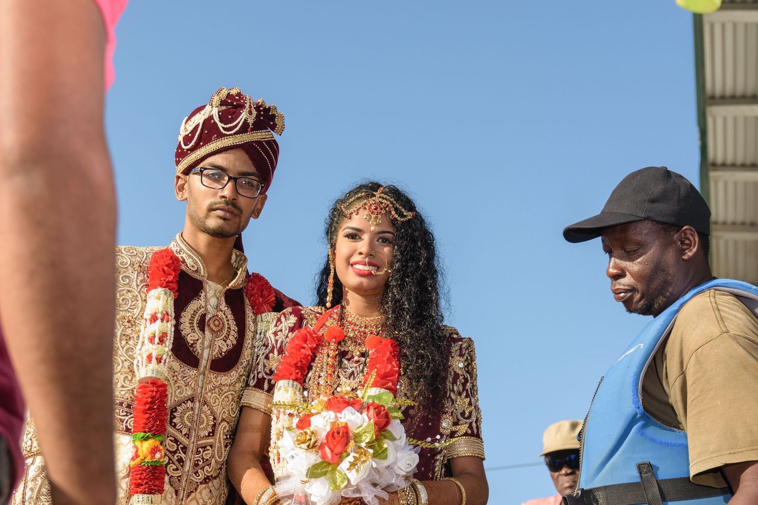 A couple in traditional Indian wedding attire stands outdoors against a blue sky.
