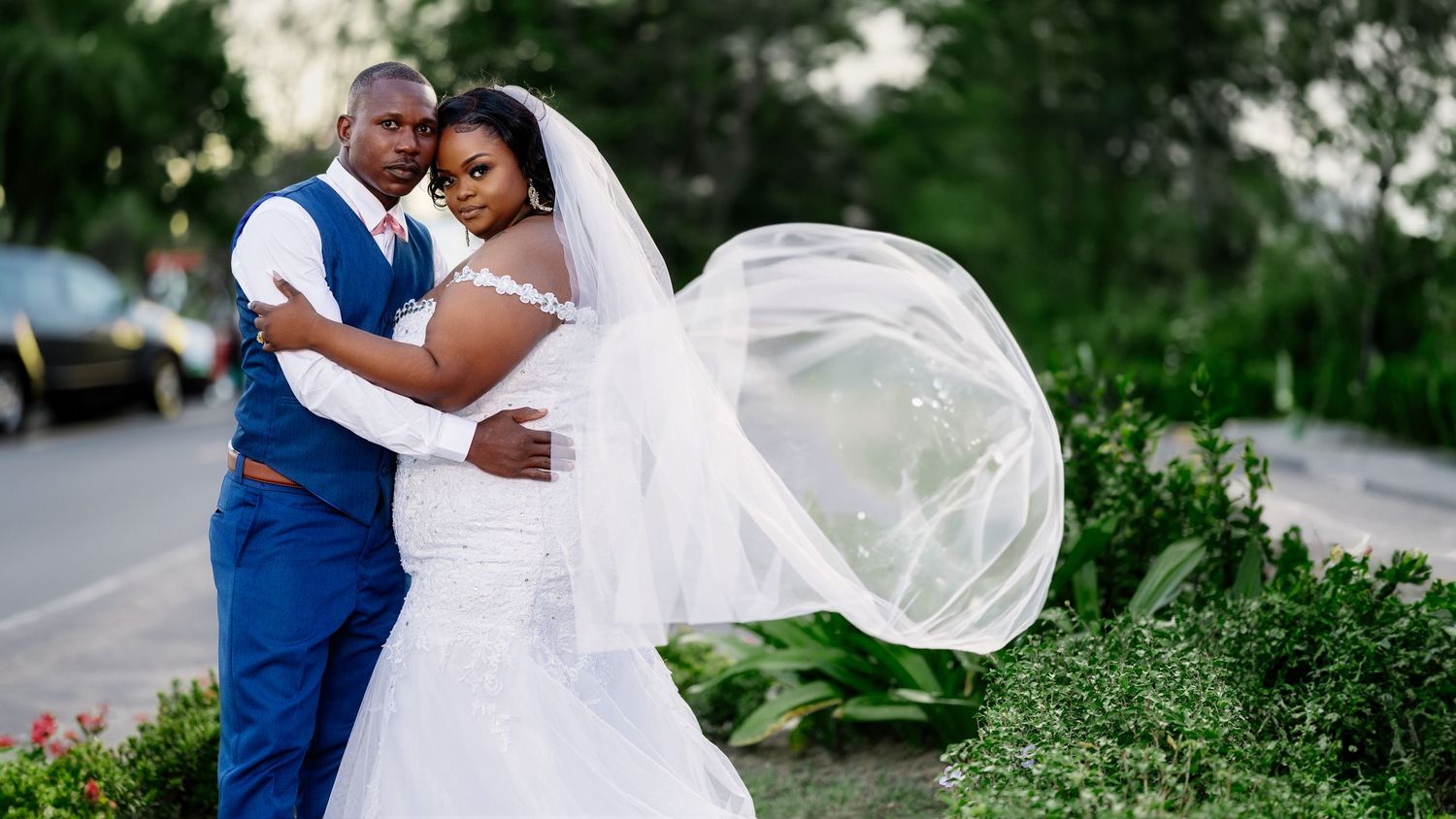 Elegant Black couple in wedding attire, bride in white gown with flowing veil, groom in blue suit, posing outdoors.