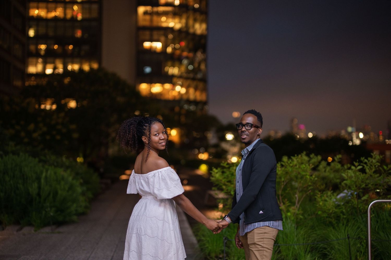 Couple shares romantic moments during golden hour photo session in an outdoor garden setting with city lights in background.