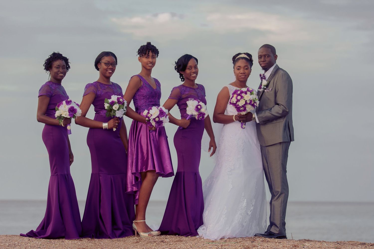 Wedding party poses on beach with bridesmaids in purple dresses and couple in formal attire.
