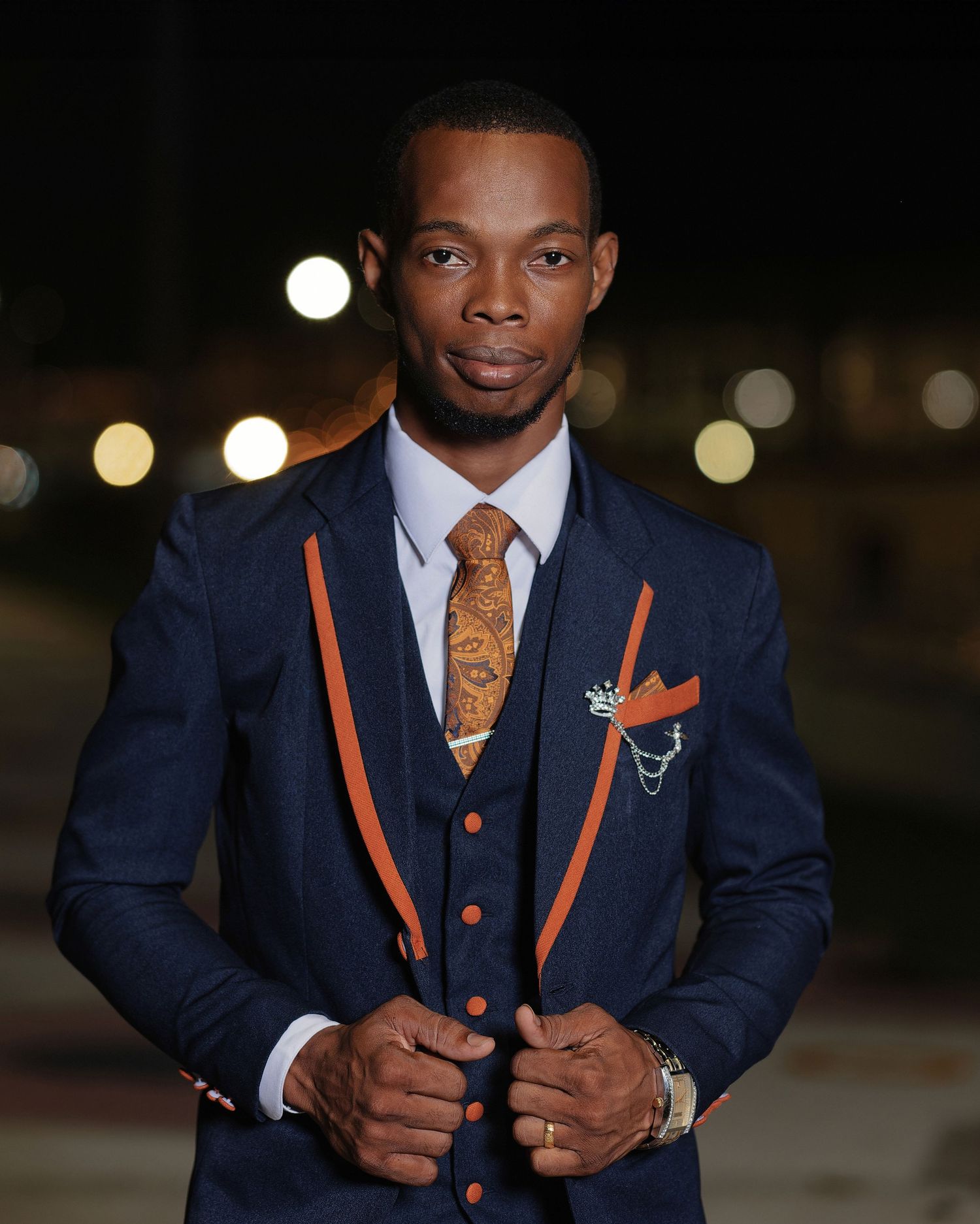 Stylish man in navy blue and orange three-piece suit with gold tie poses confidently at night.