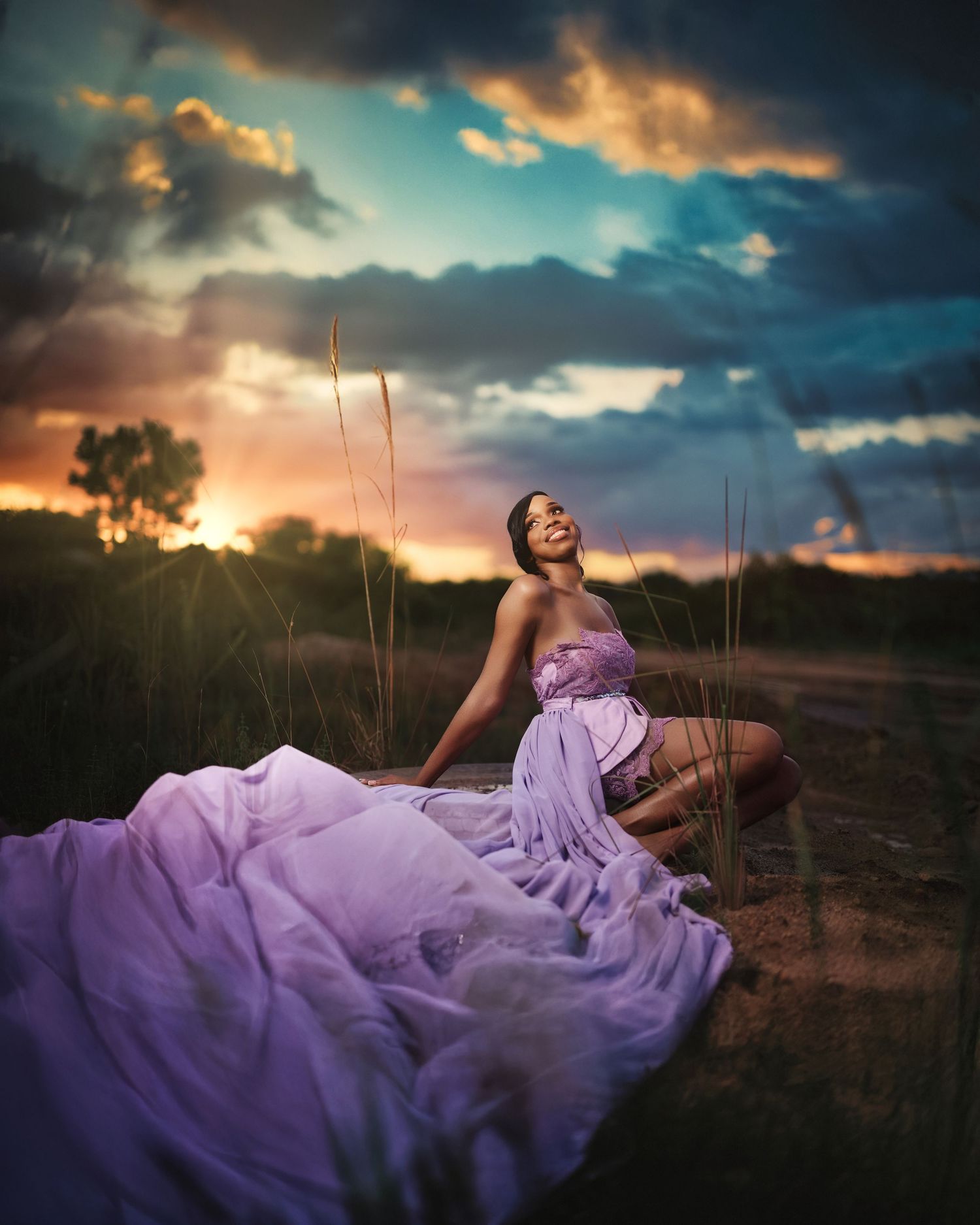 Person in flowing purple dress poses dramatically at sunset in field.