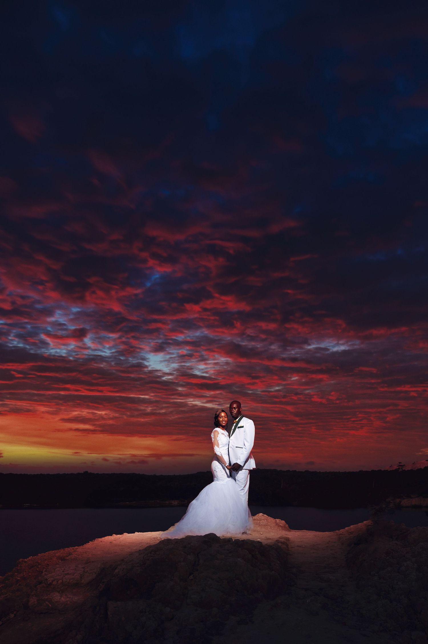 Dramatic wedding portrait against fiery red sunset clouds over water.