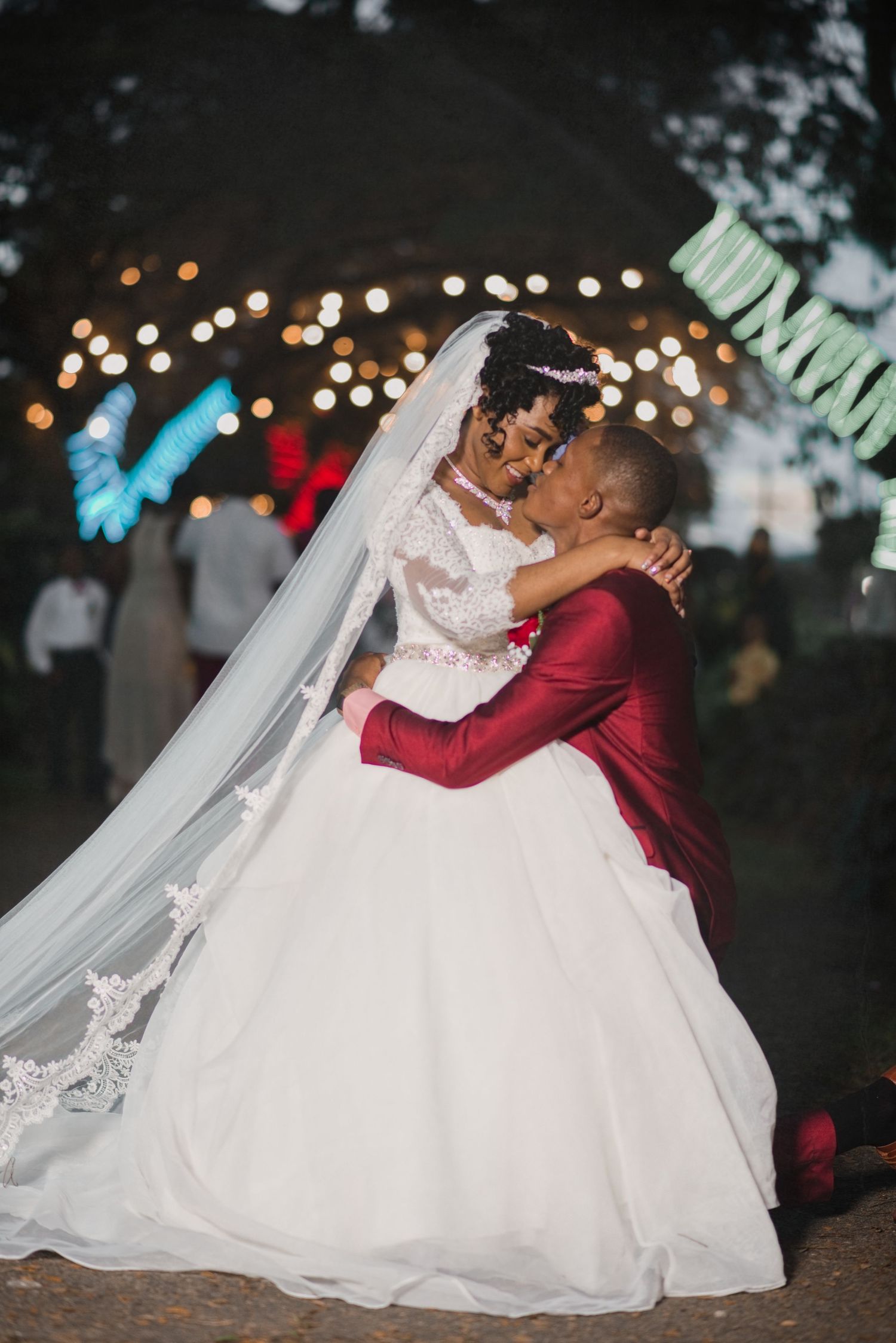 Bride and groom sharing a romantic kiss at an outdoor evening wedding reception with string lights in the background.