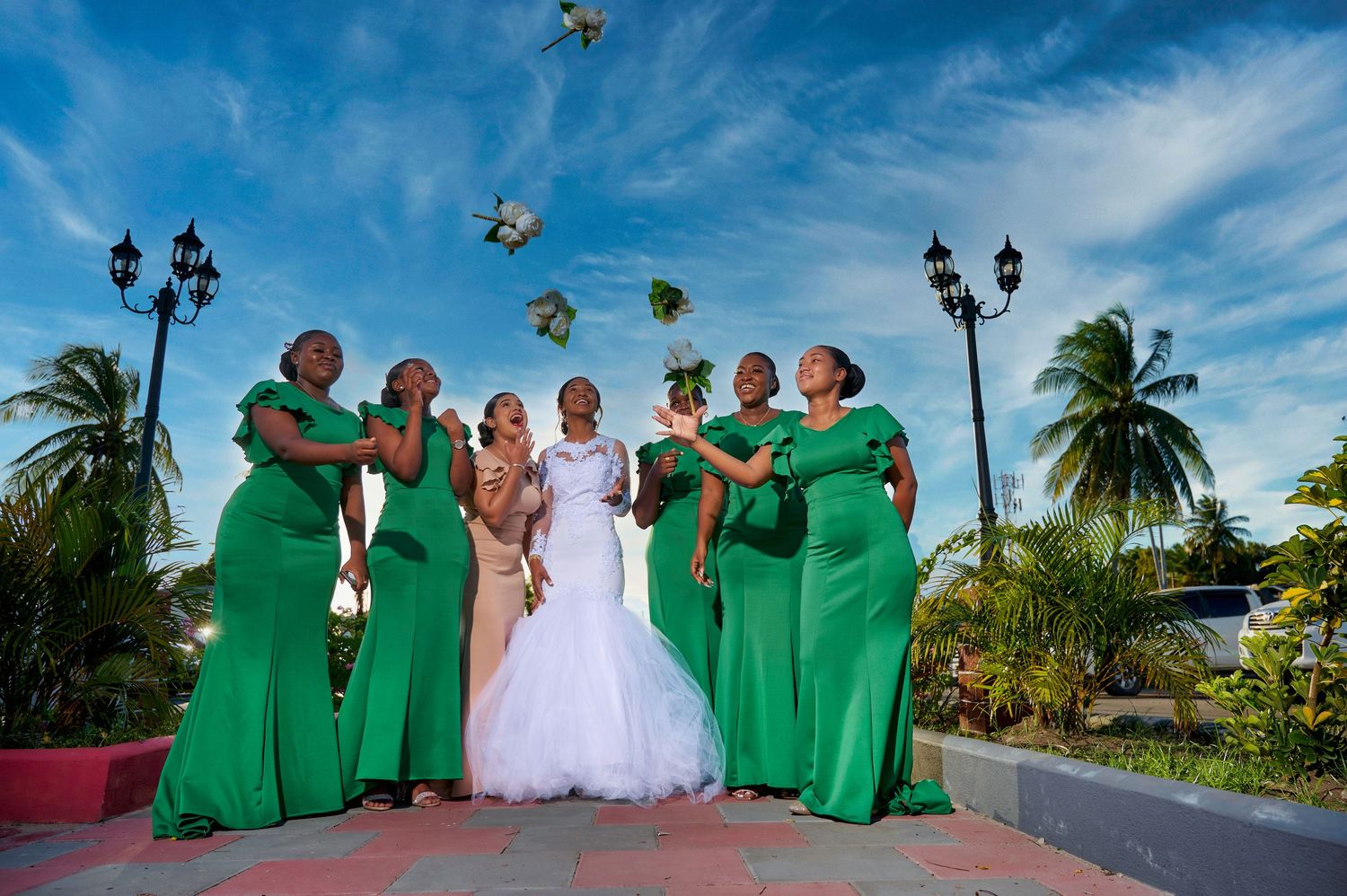 Wedding party poses outdoors with bridesmaids in emerald green dresses and bride in white gown under blue sky.