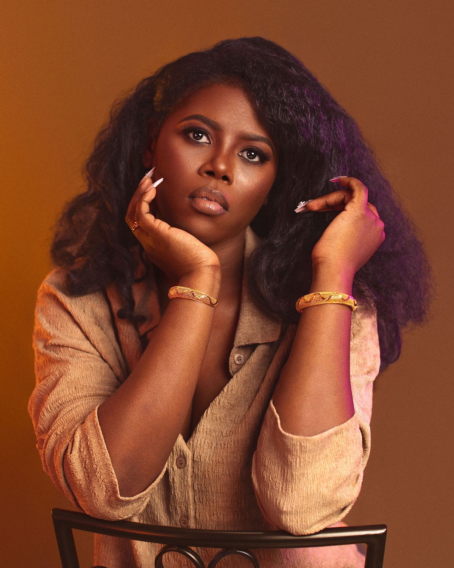A young Black woman poses gracefully with hands framing her face, wearing gold bangles and a beige blazer against a warm brown backdrop.