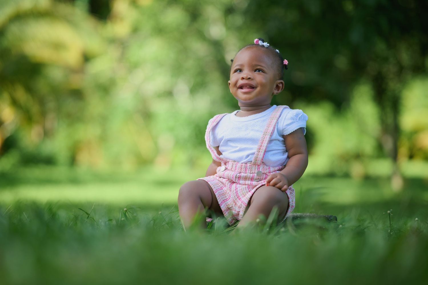 Toddler in pink and white outfit walking on grass in sunny park.