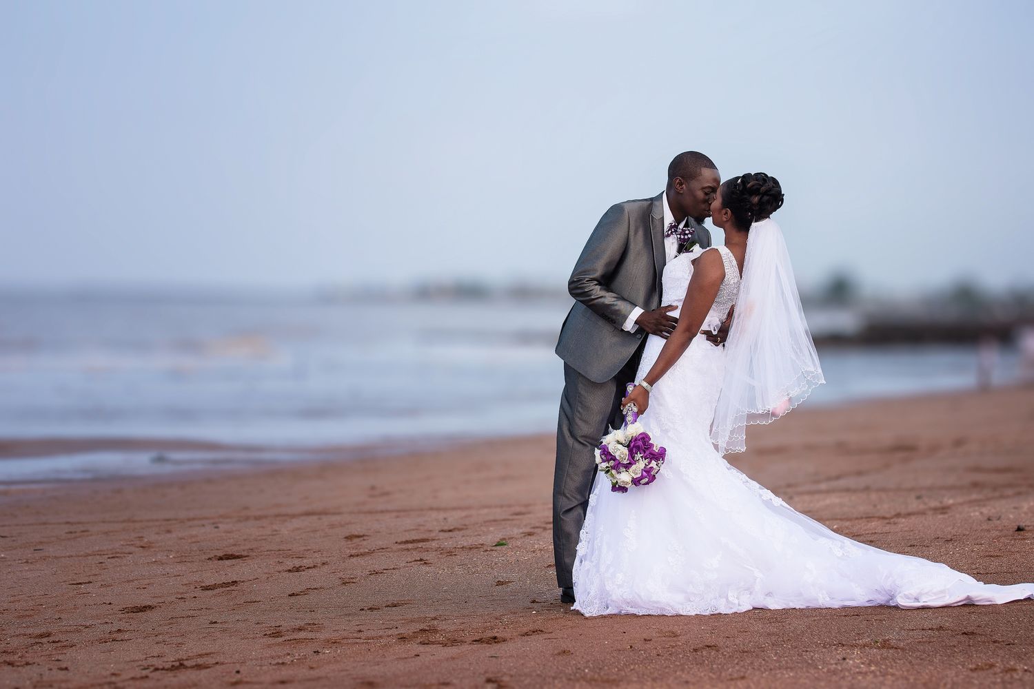 A romantic beach wedding photo of a couple in formal attire embracing by the ocean.