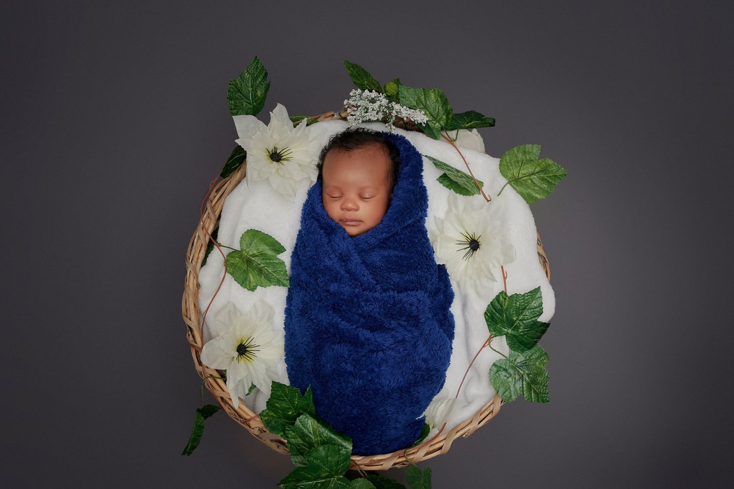 Sleeping infant in blue wrap nestled in wicker basket with white flowers.