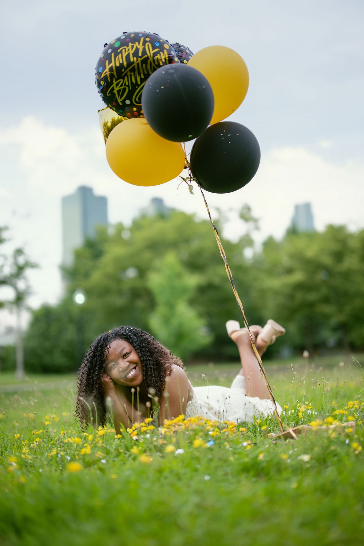 Person lying in grass holds black and yellow balloons with city skyline in background.