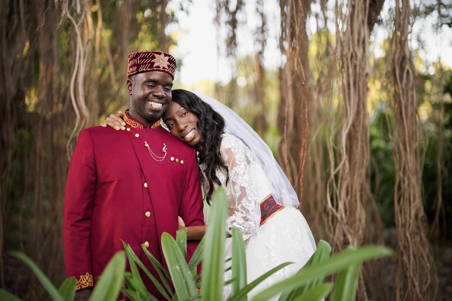 A joyful couple in traditional African wedding attire poses outdoors among lush greenery and trees.