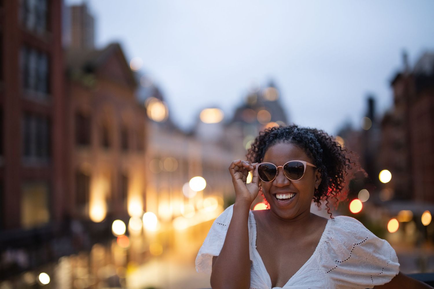 Smiling woman in white dress adjusting sunglasses outdoors at dusk with blurred city lights behind her.