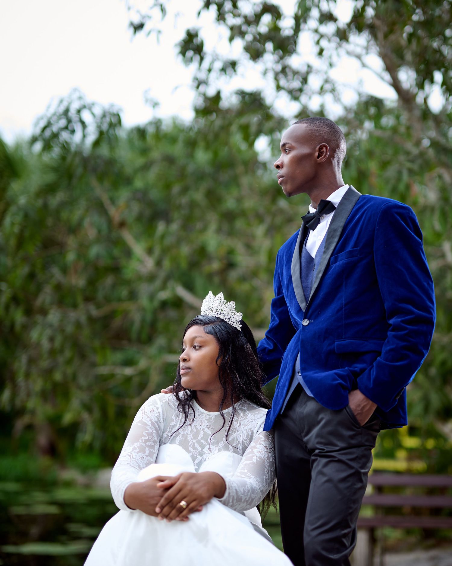 Elegant couple in wedding attire, bride in white lace gown with crown, groom in royal blue velvet tuxedo, outdoors.